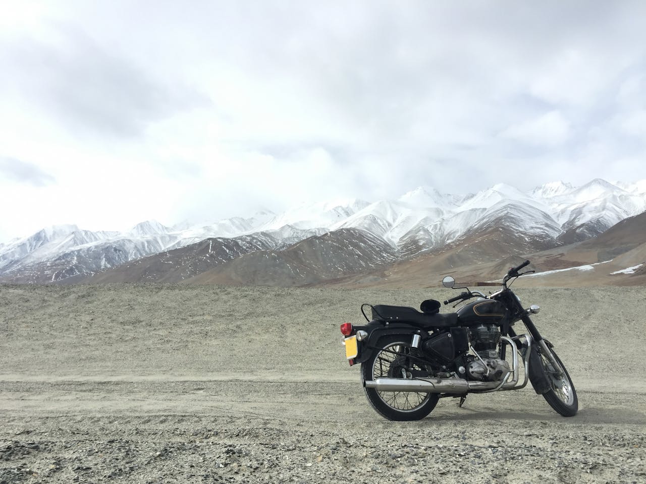 A vintage motorcycle parked on a gravel road with snowcapped mountains in the background, under a cloudy sky.